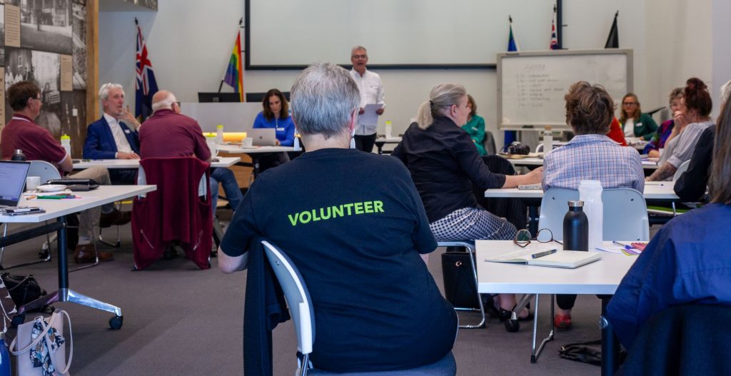 Gift of Communication participants sitting at tables discussing communication challenges with a person in a tshirt with the word volunteer on the back, featured in the foreground.